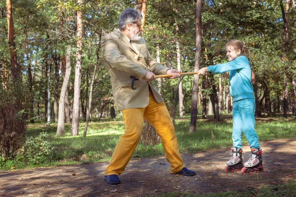 祖父は公園で孫娘と散歩する 女の子のローラーは 古い祖父と公園でスケートします 面白いおじいちゃんと小さな女の子が遊んで冗談を言っている ストックフォト