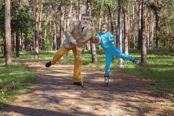 祖父は公園で孫娘と散歩する 女の子のローラーは 古い祖父と公園でスケートします 面白いおじいちゃんと小さな女の子が遊んで冗談を言っている ストック写真