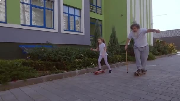 少女はスケートボードに乗るために老人に教える 彼らは路上でスケートボードに乗るように訓練しています おじいちゃんがスケートボードに乗ってる — ストック動画