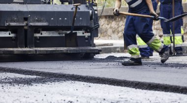 Üniformalı bir operatörün elinde sıcak asfalt olan kürek arka planda asfalt döşeme makineleri.