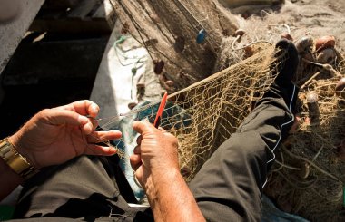 Balıkçıların elinde balık ağı, iğne ve iplikle, ayak parmaklarıyla ağı yakalıyor.