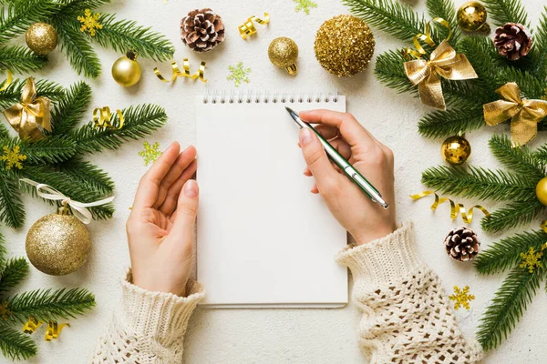 Woman writing Christmas to do list on notebook on colored background with xmas decorations.