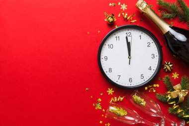 New year celebration concept with a bottle of champagne and two glasses toasting. Christmas gift box, alarm clock and fir tree branch on colored table. Top view Copy space.