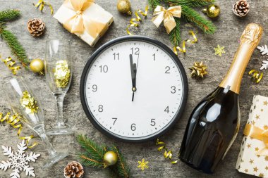 New year celebration concept with a bottle of champagne and two glasses toasting. Christmas gift box, alarm clock and fir tree branch on colored table. Top view Copy space.