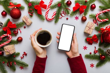 Bir kadının elinde sıcak bir fincan kahveyle akıllı bir telefonu tutması. Renkli arka planda Noel süslemesi.