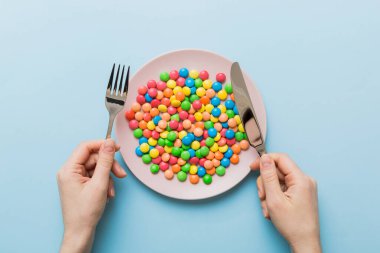 the girl holds cutlery in her hands and eats sweets in a plate. Health and obesity concept, top view on colored background.
