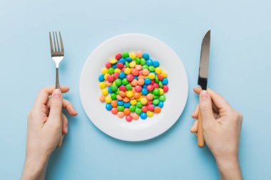 the girl holds cutlery in her hands and eats sweets in a plate. Health and obesity concept, top view on colored background.