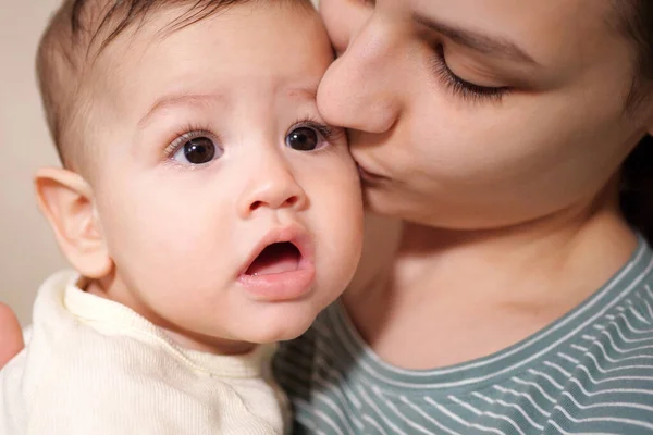 Young mother, kissing her baby boy at home. Family concept. Closeup portrait of loving mother young woman kissing little kid, home interior.