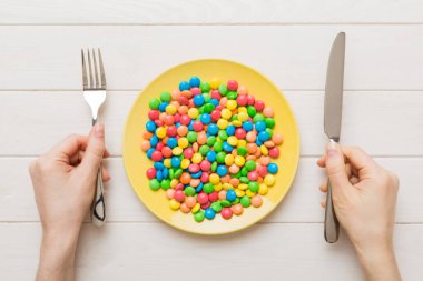 the girl holds cutlery in her hands and eats sweets in a plate. Health and obesity concept, top view on colored background.