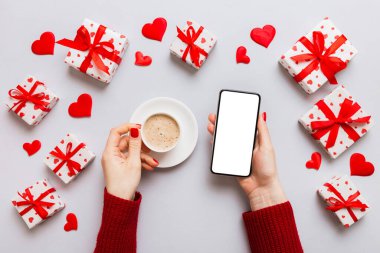 Female hands holding smart phone with coffee for Valentine day, gift box and envelope, hearts.