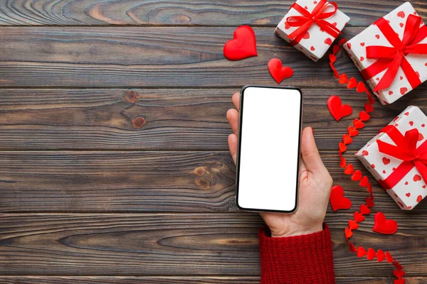 Woman hand holding mobile phone with blank screen on colored background with hearts, valentine day concept top view flat lay.