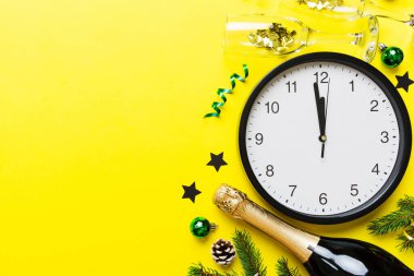 New year celebration concept with a bottle of champagne and two glasses toasting. Christmas gift box, alarm clock and fir tree branch on colored table. Top view Copy space.