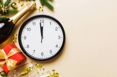 New year celebration concept with a bottle of champagne and two glasses toasting. Christmas gift box, alarm clock and fir tree branch on colored table. Top view Copy space.