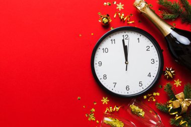 New year celebration concept with a bottle of champagne and two glasses toasting. Christmas gift box, alarm clock and fir tree branch on colored table. Top view Copy space.