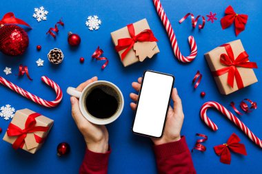 Bir kadının elinde sıcak bir fincan kahveyle akıllı bir telefonu tutması. Renkli arka planda Noel süslemesi.