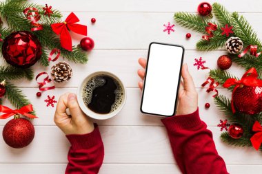 Bir kadının elinde sıcak bir fincan kahveyle akıllı bir telefonu tutması. Renkli arka planda Noel süslemesi.