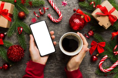 Bir kadının elinde sıcak bir fincan kahveyle akıllı bir telefonu tutması. Renkli arka planda Noel süslemesi.