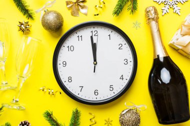 New year celebration concept with a bottle of champagne and two glasses toasting. Christmas gift box, alarm clock and fir tree branch on colored table. Top view Copy space.