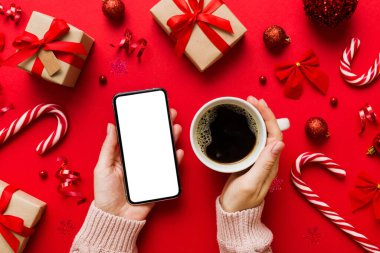 Bir kadının elinde sıcak bir fincan kahveyle akıllı bir telefonu tutması. Renkli arka planda Noel süslemesi.
