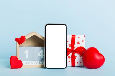 mobile phone with blank screen on colored background with hearts, calendar and gift box, valentine day 14 february concept perspertive view flat lay.