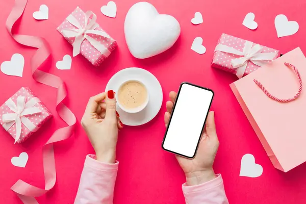 Female hands holding smart phone with coffee for Valentine day, gift box and envelope, hearts.