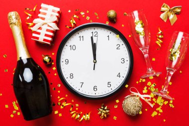 New year celebration concept with a bottle of champagne and two glasses toasting. Christmas gift box, alarm clock and fir tree branch on colored table. Top view Copy space.