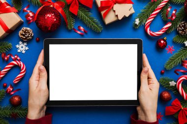 Evden Noel alışverişi, kadın elleri boş beyaz ekranlı tableti tutuyor. Kadın elinde tablet, boş ekran, Noel ağacı ve arka planda hediyeler..