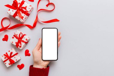 Woman hand holding mobile phone with blank screen on colored background with hearts, valentine day concept top view flat lay.