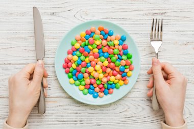 the girl holds cutlery in her hands and eats sweets in a plate. Health and obesity concept, top view on colored background.
