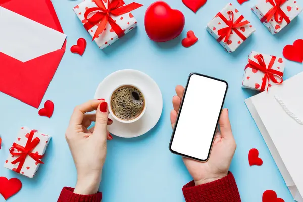 Female hands holding smart phone with coffee for Valentine day, gift box and envelope, hearts.