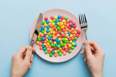 the girl holds cutlery in her hands and eats sweets in a plate. Health and obesity concept, top view on colored background.