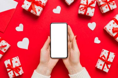 Woman hand holding mobile phone with blank screen on colored background with hearts, valentine day concept top view flat lay.