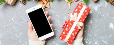 Bir elinde telefon, diğerinde de çimento arka planda hediye tutan bir kadının üst görüntüsü. Firavun ağacı ve tatil süslemeleri. Noel tatili kavramı. Model.