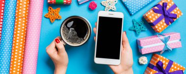 Bir elinde telefon, diğer elinde kahve tutan bir kadının mavi arka plandaki görüntüsü. Noel süslemeleri ve oyuncakları. Yeni yıl tatili konsepti. Model.