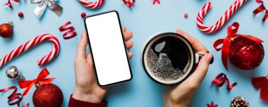 Bir kadının elinde sıcak bir fincan kahveyle akıllı bir telefonu tutması. Renkli arka planda Noel süslemesi.