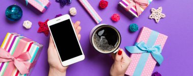 Bir elinde telefon, diğer elinde bir fincan kahve olan bir kadının mor arka planda görüntüsü. Noel süslemeleri ve oyuncakları. Yeni yıl tatili konsepti. Model.