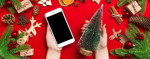 Bir elinde telefon, diğer elinde de kırmızı arka planda Noel ağacı tutan kadınların üst görüntüsü. Yeni yıl tatili konsepti. Model.