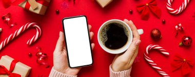 Bir kadının elinde sıcak bir fincan kahveyle akıllı bir telefonu tutması. Renkli arka planda Noel süslemesi.
