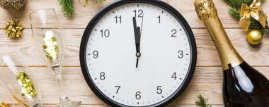 New year celebration concept with a bottle of champagne and two glasses toasting. Christmas gift box, alarm clock and fir tree branch on colored table. Top view Copy space.