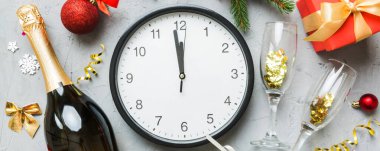 New year celebration concept with a bottle of champagne and two glasses toasting. Christmas gift box, alarm clock and fir tree branch on colored table. Top view Copy space.