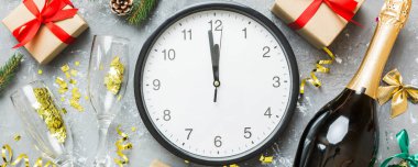 New year celebration concept with a bottle of champagne and two glasses toasting. Christmas gift box, alarm clock and fir tree branch on colored table. Top view Copy space.