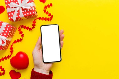 Woman hand holding mobile phone with blank screen on colored background with hearts, valentine day concept top view flat lay.