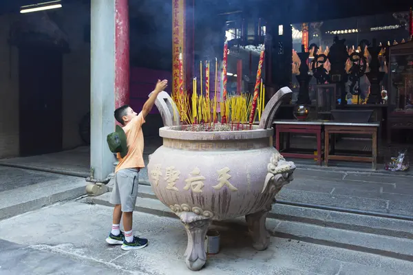 HO CHI MINH CITY, VİETNAM, 17 Ağustos 2024: Genç bir çocuk Vietnam 'daki geleneksel bir tapınakta yanan yosun çubuklarını adak olarak tutuyor, büyük bir tütsü vazosundan dumanlar yükseliyor.