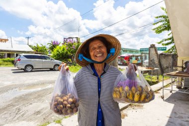 HO CHI MINH CITY, VİETNAM, 18 Ağustos 2024: Geleneksel konik bir şapka takan neşeli bir Vietnamlı kadın, güneşli bir sokakta satılık uzun ve muz torbaları tutarken gülümsüyor