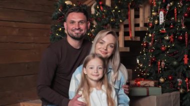 Üç kişilik mutlu bir aile, baba, anne ve kız, evdeki Noel ağacının yanında bir aile fotoğrafı için poz veriyorlar. Evli çift, yanıp sönen ışıkların yanında sarılıyor. Bayram havası esiyor.