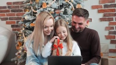 Üç kişilik genç bir aile, baba, anne ve kız Noel ağacının yanında oturmuş dizüstü bilgisayarla bir video görüşmesi yapıyorlar.