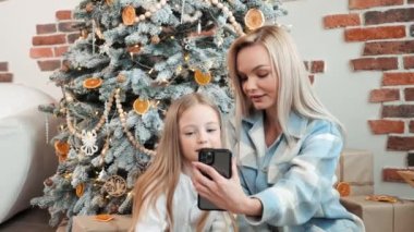 Mutlu anne ve kız günlük giysiler içinde cep telefonuyla video görüşmesi yapıyor, Noel ağacı, şenlik ve iletişim teknolojisi yakınlarındaki arkadaşlarını veya akrabalarını gösteriyor. Yeni yıl. Tatil
