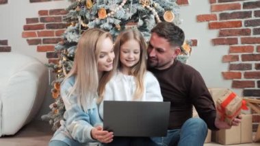 Üç kişilik şirin mutlu bir aile, baba, anne ve kız Noel ağacının yanında oturmuş dizüstü bilgisayarla bir video görüşmesi yapıyorlar, çocuk konferansta arkadaşlarına hediye sunuyor.