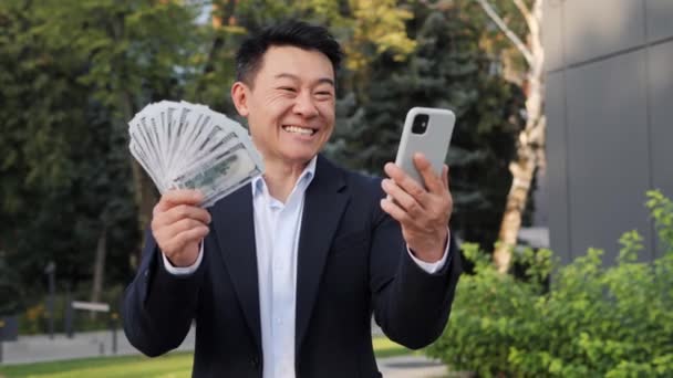 形象滑稽快乐的韩国商人挥挥手 在街上拿着手机跳舞 成功的商人看上去对外面的美元感到满意 庆祝胜利 — 图库视频影像