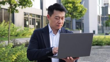Uzaktan kumandayla çalışan Asyalı odaklı iş adamı bilgisayarlı taşınabilir dizüstü bilgisayar çevrimiçi sohbet sohbet programcısı uygulaması ofis bilişim programlama şirketinin yanında duruyor, konsantre erkek yüzü